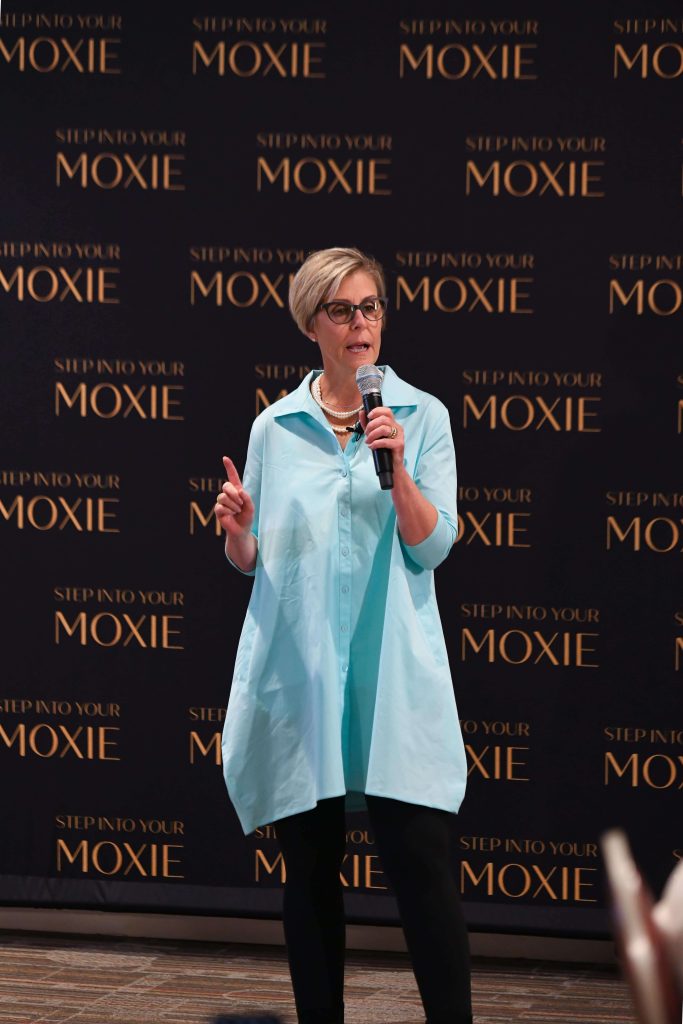 Kim Boudreau Smith with microphone, on stage and speaking with a Step into your Moxie background